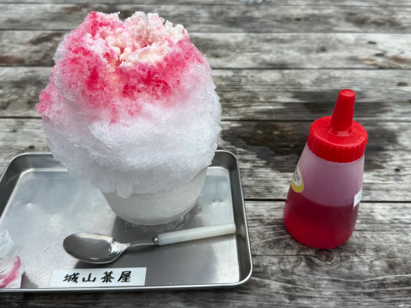 A towering mound of shaved ice with red syrup on a metal tray labelled Shiroyama Chaya, next to a squeeze bottle of extra syrup