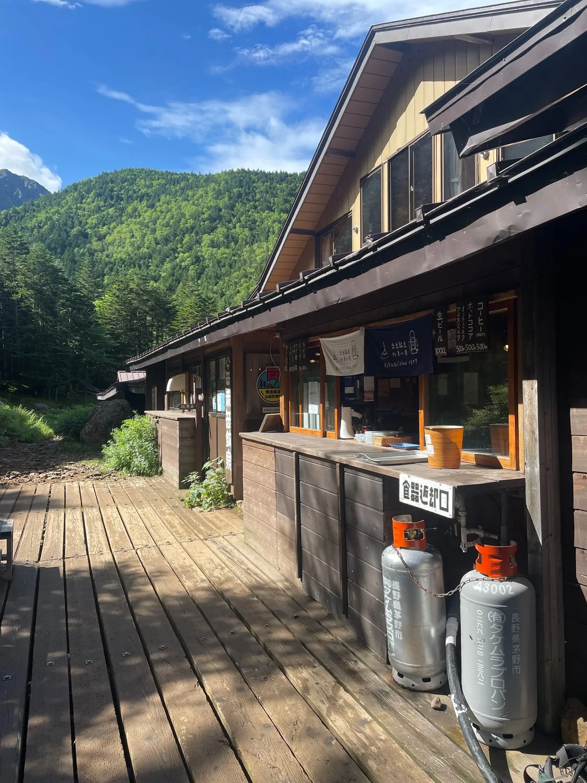 Akadake Kosen, a wooden mountain hut in Yatsugatake, on a sunny summer morning, with a wooden deck, gas cylinders and a forested ridge behind